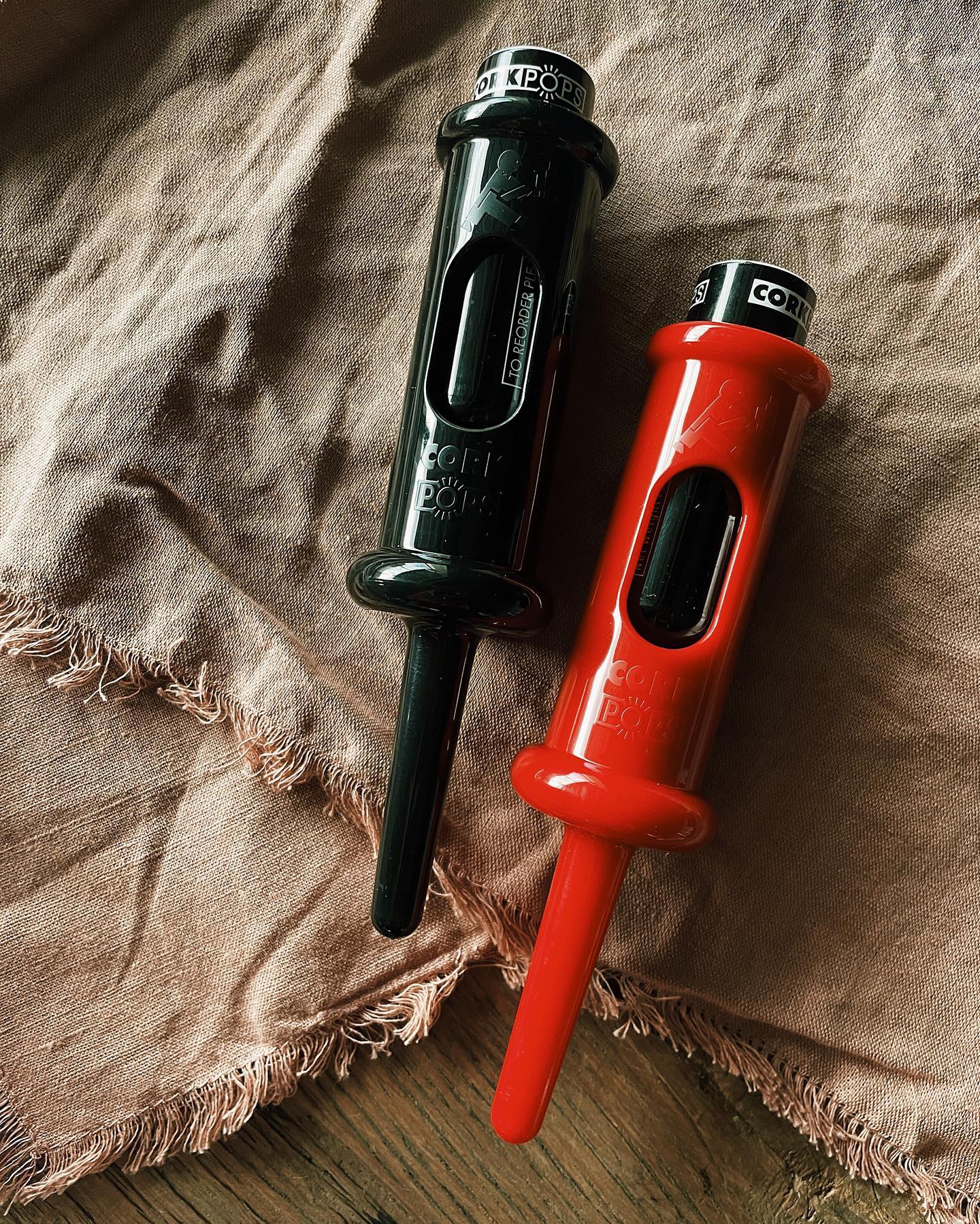 Two Original Cork Pops openers in black and red resting on linen