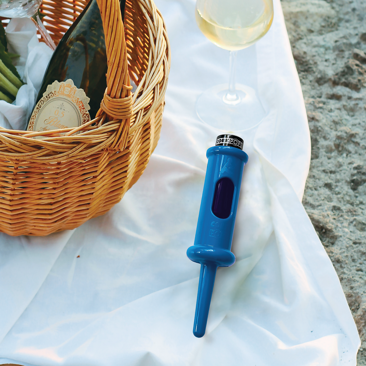 Ocean blue Cork Pops opener on a white cloth beside a picnic basket